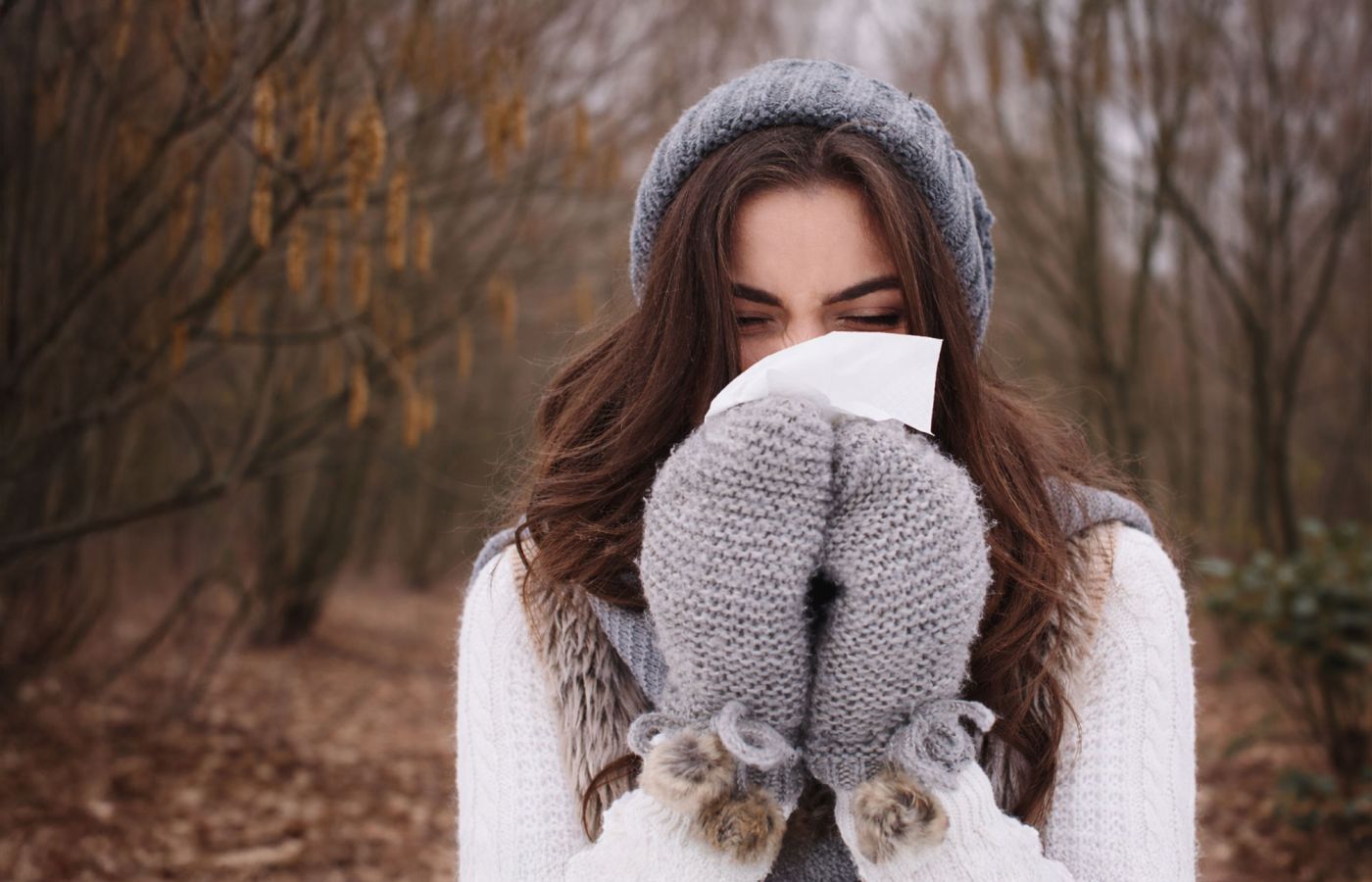 Frau mit Mütze und Fäustlingen hält ein Taschentuch vor Nase und Mund im Wald.